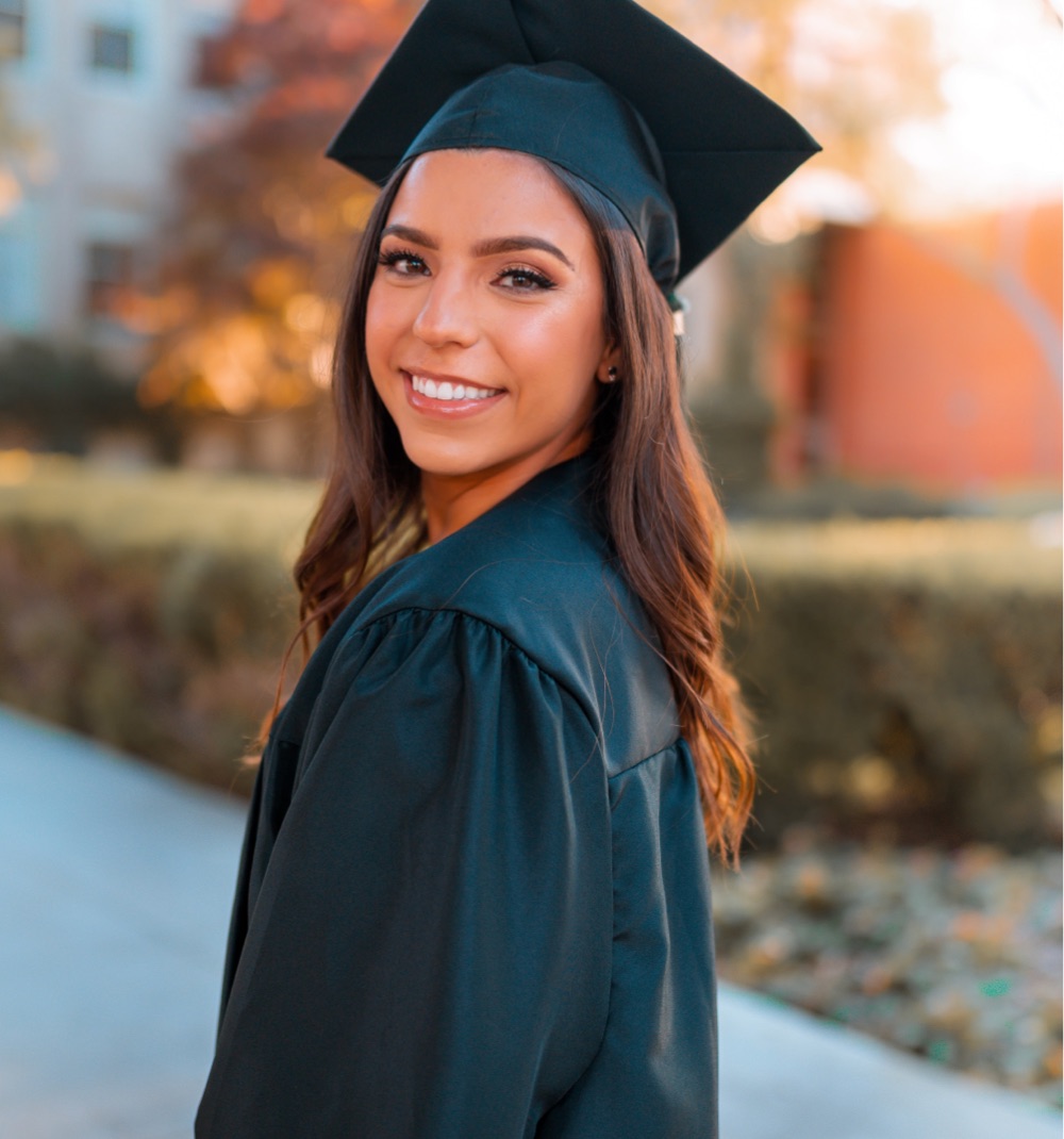 Young woman wearing a graduation cap and gown looking over her shoulder at camera
